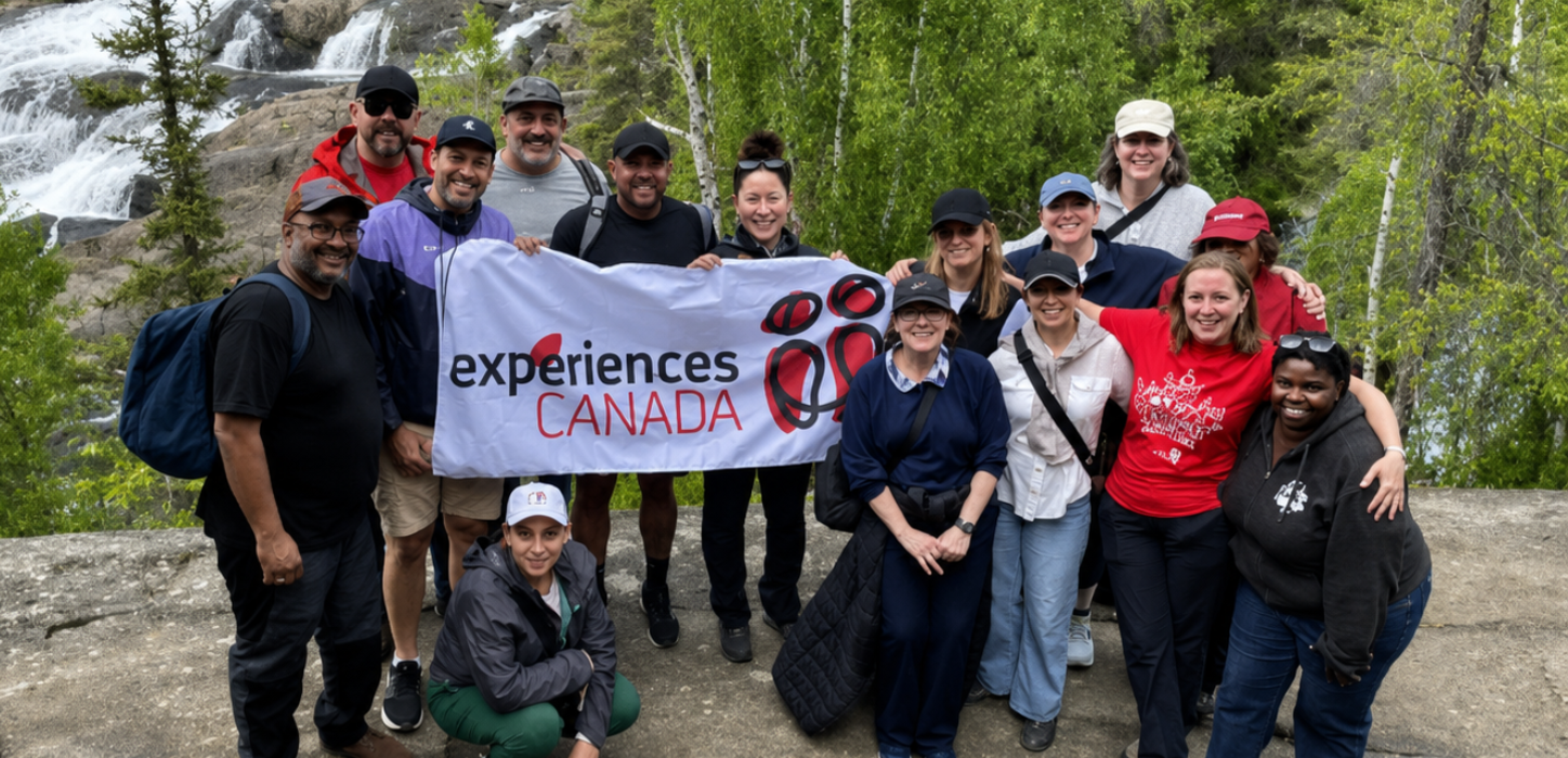 A group of Experiences Canada staff members pose together on a rocky overlook, holding an Experiences Canada banner in front of a scenic waterfall surrounded by lush green trees.